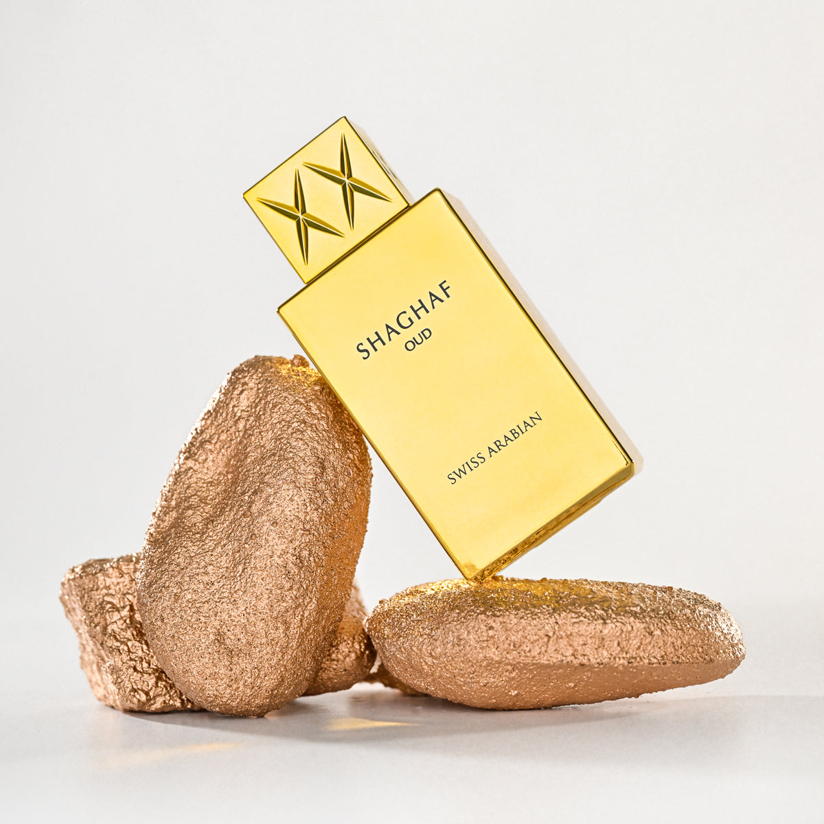 Gold Shaghaf Oud perfume bottle with two gold rocks on a light gray background