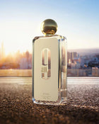 The image shows an elegant, clear glass perfume bottle with a reflective gold cap, placed on a textured surface against an out-of-focus cityscape bathed in the warm, golden light of sunrise or sunset. Further details at the bottom of the label read "eau de parfum vaporisateur," "100ml," and "3.4fl.oz." The lighting gives the scene a luxurious and aspirational quality. 