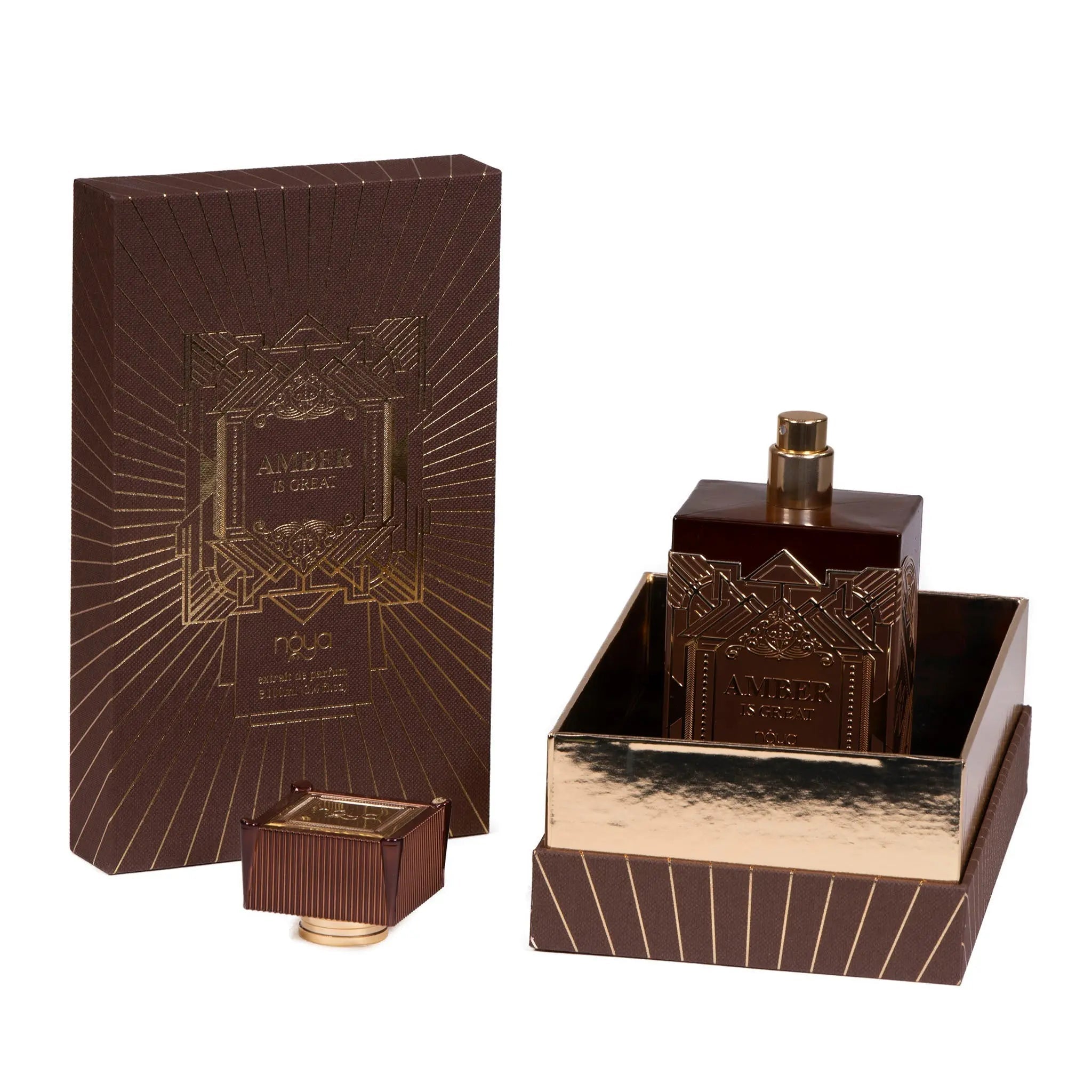 A perfume packaging and bottle set for "AMBER IS GREAT" by náyá. The packaging is a dark brown box with gold geometric patterns and lines, featuring the brand name and perfume title in gold lettering. The square glass bottle has a dark brown color with a golden cap and is adorned with the same geometric pattern as the box. The bottle is placed in an open drawer-style compartment in the box, which has a reflective gold interior.