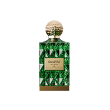 A luxurious bottle of Emerald Oud perfume by Ibraheem Al-Qurashi, featuring a geometric emerald green glass design with reflective triangular facets. The bottle is topped with a textured, spherical gold cap and has a gold nameplate on the front engraved with 'Emerald Oud' in elegant script along with Arabic text and branding. A solid gold base completes the opulent look, conveying a rich and sophisticated aesthetic.