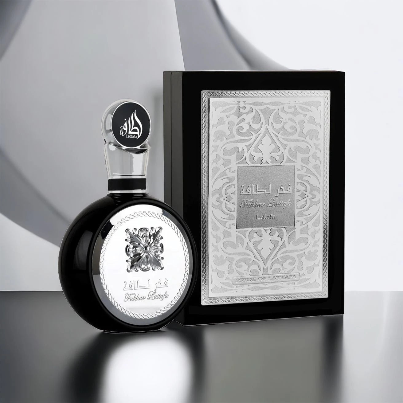 An image of a black and silver perfume bottle alongside its packaging. The bottle features a reflective black surface with a silver flower emblem and a silver cap with Arabic calligraphy. The box is a monochrome grey with elegant arabesque designs and the name "Fakhar Lattafa" in the center, along with "PRIDE OF LATTAFA" below it.