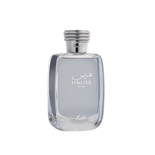 A close-up of the HAWAS For Him perfume bottle by Al Rasasi. It's a clear, rectangular glass bottle with rounded edges, filled with a light blue tinted liquid. The bottle features elegant white Arabic and English lettering that reads "HAWAS For Him." The cap is a textured silver, resembling a coiled rope design, which adds a sophisticated touch. The base of the bottle also has a delicate, white Arabic script logo.