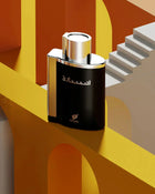 The image shows a modern and stylish perfume bottle placed in an abstract geometric setting. The bottle has a glossy black finish with a silver cap, and it is rectangular with rounded edges.  The bottle is positioned against a visually striking background composed of bold geometric shapes and lines in shades of yellow and orange, creating a three-dimensional effect that resembles architectural structures and staircases.