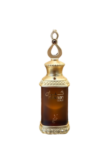 This image features a perfume bottle labeled "Kayaan Gold" resting on a glass surface. The bottle has a regal design with a gold-toned cap adorned with decorative elements and a loop on top, suggesting it can be used as a pendant or for easy carrying. The dark amber liquid inside indicates the perfume might have a rich and possibly woody or oriental fragrance.