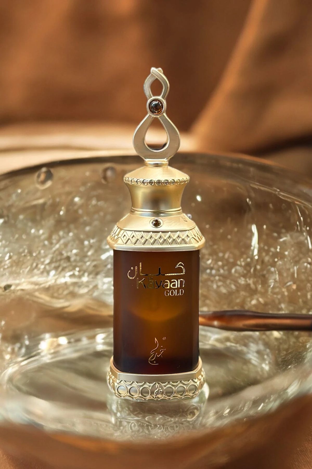 This image features a perfume bottle labeled "Kayaan Gold" resting on a glass surface. The bottle has a regal design with a gold-toned cap adorned with decorative elements and a loop on top, suggesting it can be used as a pendant or for easy carrying. The dark amber liquid inside indicates the perfume might have a rich and possibly woody or oriental fragrance.