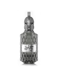 A product shot of Le Falcone Niche Collection’s “Khulood” perfume bottle on a white background: a tall, amber‑colored glass vessel with a diamond‑cut texture and a polished bronze plaque engraved with Arabic calligraphy above the word “Khulood.” The bottle is crowned by an ornate bronze cap sculpted into a stylized warrior’s helmet with a crest‑style loop at the top, conveying a luxurious, sculptural aesthetic.