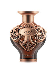 A high‑resolution product shot of Le Falcone Niche Collection’s “Turath Intense” perfume bottle against a white background. The bottle has a dark amber, heart‑shaped glass body topped with a wide, copper‑toned neck and cap. Its upper half is covered in ornate, scrolling filigree in a polished copper finish, forming elegant arabesque patterns. A matching copper plaque at the front bears Arabic calligraphy above the word “TURATH,” and “INTENSE” appears in gold lettering near the base.