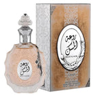 The image features a perfume bottle and its packaging:  The perfume bottle is crafted from multi-faceted glass with a pale pink tint and a decorative surface texture. It is adorned with a clear glass stopper that has a detailed pattern and a metallic chain with a charm attached to the neck. The label on the bottle has Arabic calligraphy and the name "ROUAT AL MUSK" in English.