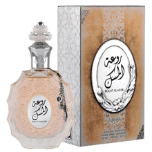 The image features a perfume bottle and its packaging:  The perfume bottle is crafted from multi-faceted glass with a pale pink tint and a decorative surface texture. It is adorned with a clear glass stopper that has a detailed pattern and a metallic chain with a charm attached to the neck. The label on the bottle has Arabic calligraphy and the name "ROUAT AL MUSK" in English.