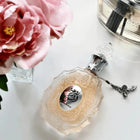 The image features a perfume bottle on a white surface:  The bottle is crafted from clear glass with a textured pattern and a pale pink tint. It has an ornate clear glass stopper and a metallic chain with a charm attached to the neck. A label with Arabic calligraphy and the name "ROUAT AL MUSK" in English is displayed on the front.