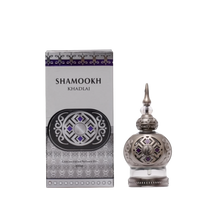 The image shows a product called "Shamookh" by Khadlaj, which is a concentrated perfume oil. The perfume comes in a 20ml spherical bottle with a silver finish, featuring intricate patterns and a pointed cap that adds a touch of elegance. The design includes decorative elements with purple accents, enhancing its luxurious appearance. The packaging box matches the bottle's design, with a silver and purple theme, featuring the product name "Shamookh" in bold letters and additional ornamental details.