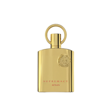 The image shows a bottle of "Supremacy" eau de parfum by Afnan Perfumes. The bottle is sleek and made of reflective gold metal, giving it a luxurious and elegant appearance. The design features a circular ring attached to the top, which serves as the cap. The front of the bottle is adorned with a raised emblem featuring intricate details, positioned near the top right corner.