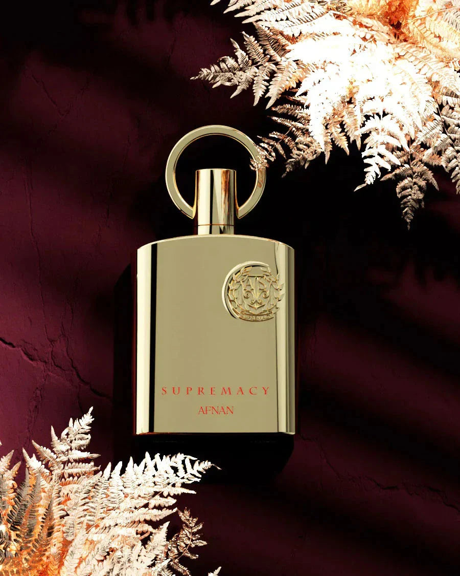 The image features an elegant, matte gold perfume bottle with a sleek design, positioned in front of a rich, dark maroon backdrop with metallic gold fern leaves radiating in the corner, creating a luxurious and opulent atmosphere. Below the emblem, the words "SUPREMACY AFNAN" are printed in a refined, understated font, indicating the brand and perhaps the fragrance line. The overall composition of the image exudes a feeling of premium quality and exclusivity. 
