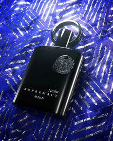The image features an elegant black perfume bottle with a reflective, ring-shaped cap. The bottle is emblazoned with a silver emblem and the text "NOIR SUPREMACY AFNAN" in a sophisticated white font. It is placed against a vivid blue background adorned with a pattern of shiny, tropical palm leaves. The contrast between the dark bottle and the bright, luminescent blue creates a striking visual presentation, highlighting the product's luxurious appeal.
