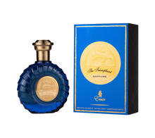 A round, deep-blue perfume bottle with a ridged design and a gold-toned cap, featuring a circular gold emblem depicting a ship on the front. It is displayed alongside a coordinating royal-blue rectangular box with gold trim and the same ship emblem, labeled “The Triumphant Sapphire” by Emir (100 ml Eau De Parfum).