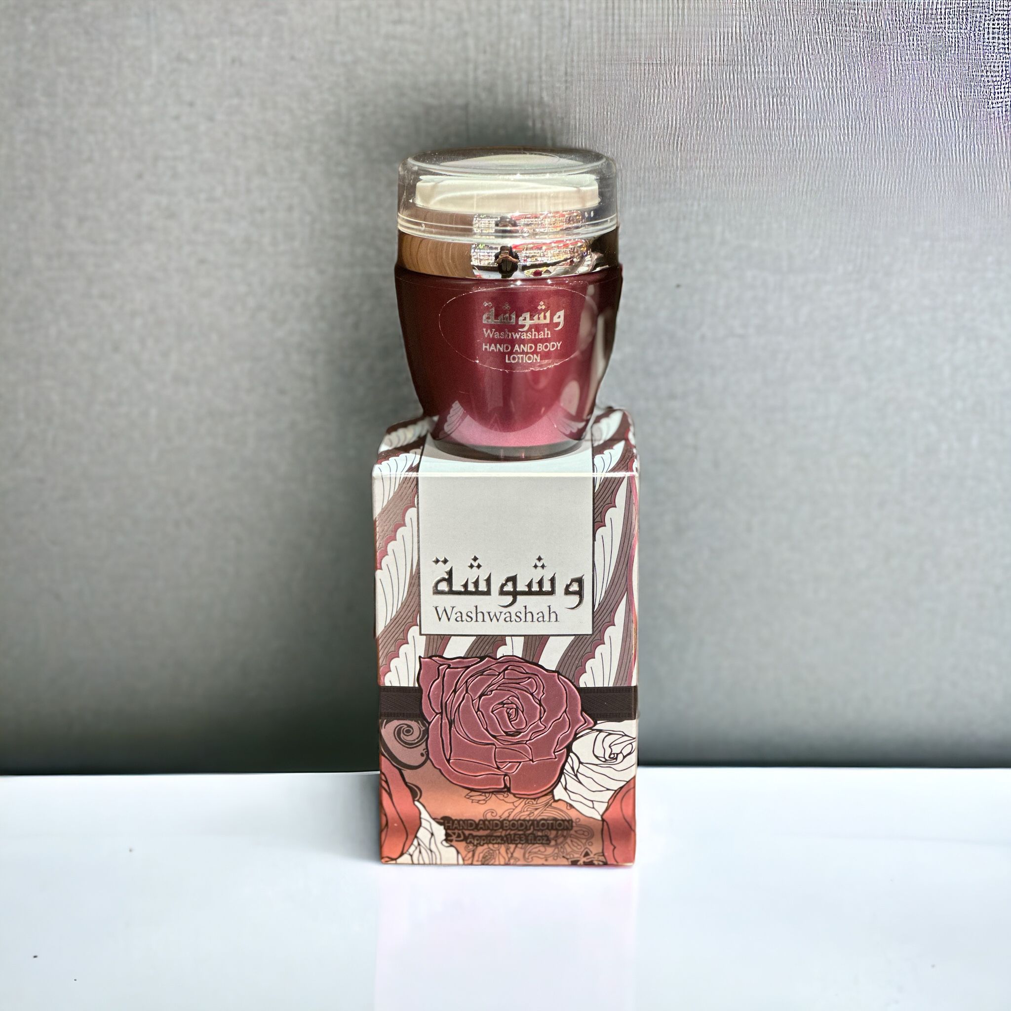 The image shows a product named "Washwashah Hand and Body Lotion." The lotion comes in a dark red container with a clear lid and is placed on top of a matching red and beige box adorned with intricate floral patterns and Arabic calligraphy. The box prominently features the product name "Washwashah" along with an illustration of a rose. The elegant and artistic packaging suggests a luxurious and high-quality skincare product.
