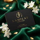 The black "LAMILAS PERFUME" gift card with gold lettering is placed on emerald green velvet, surrounded by white jasmine flowers and small gold leaves.