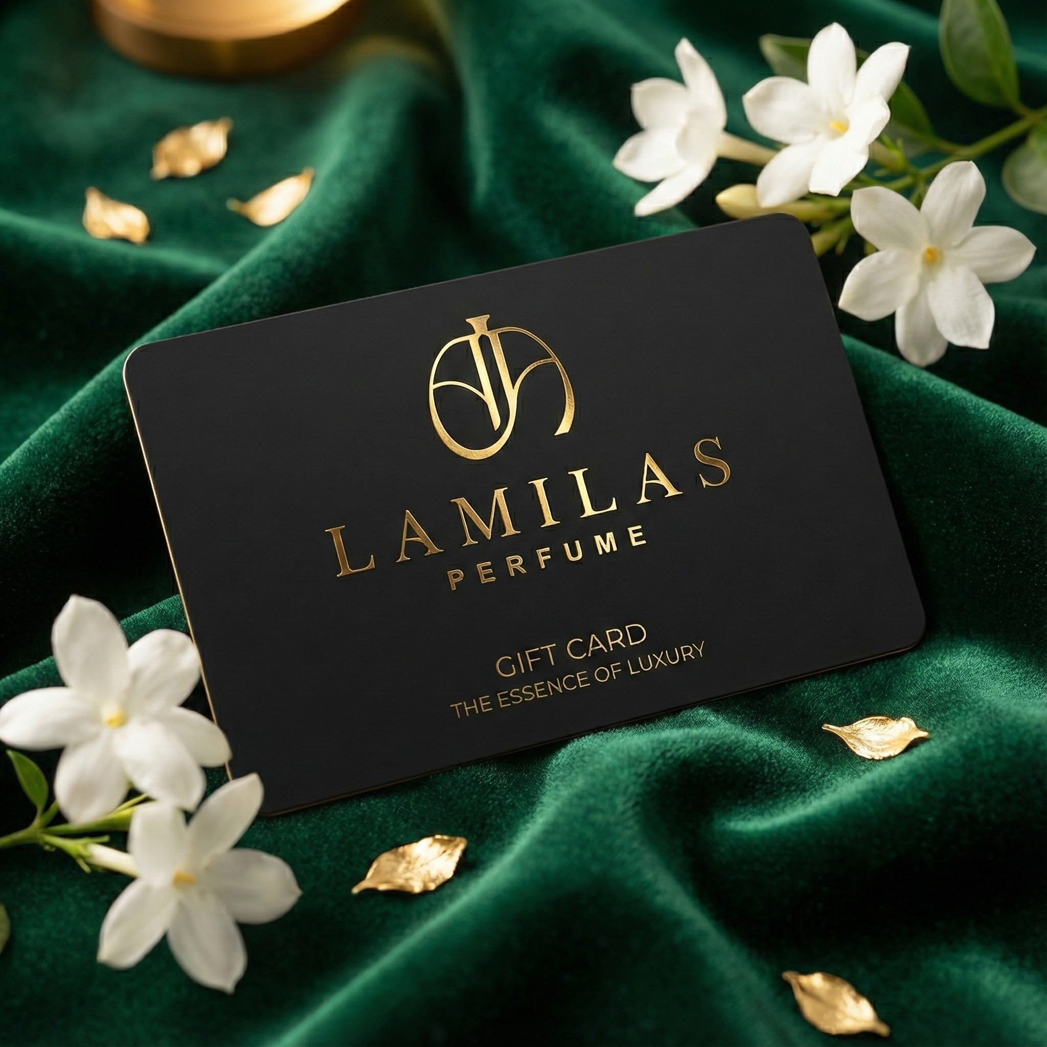 The black "LAMILAS PERFUME" gift card with gold lettering is placed on emerald green velvet, surrounded by white jasmine flowers and small gold leaves.