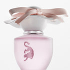A frosted light pink perfume bottle with a matching pink leopard emblem on the front, topped with a white cap and a large pale pink satin bow, shown on a clean white background.